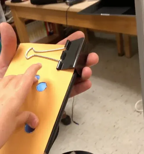 A hand resting on a cardstock overlay clipped to a tablet with a binder clip. Holes cut in the card show the blue touchscreen beneath each fingertip.